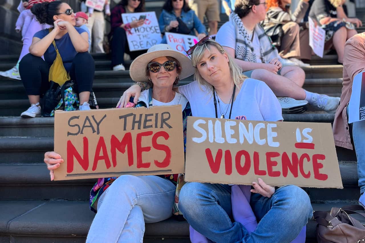 Two women sit on steps holding signs that read "SAY THEIR NAMES" and "SILENCE IS VIOLENCE" during a protest.