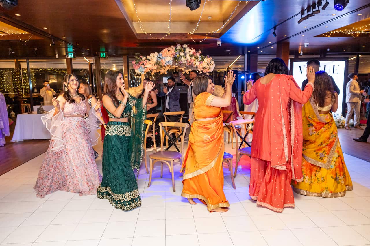 Several women in colourful Indian attire are dance around empty chairs on a white tiled floor beneath a floral archway.