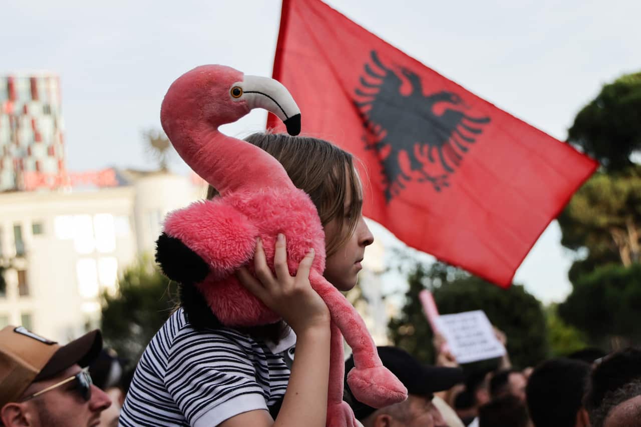 A young girl holds a stuffed flamingo in a crowd as an Albanian flag waves behind her.