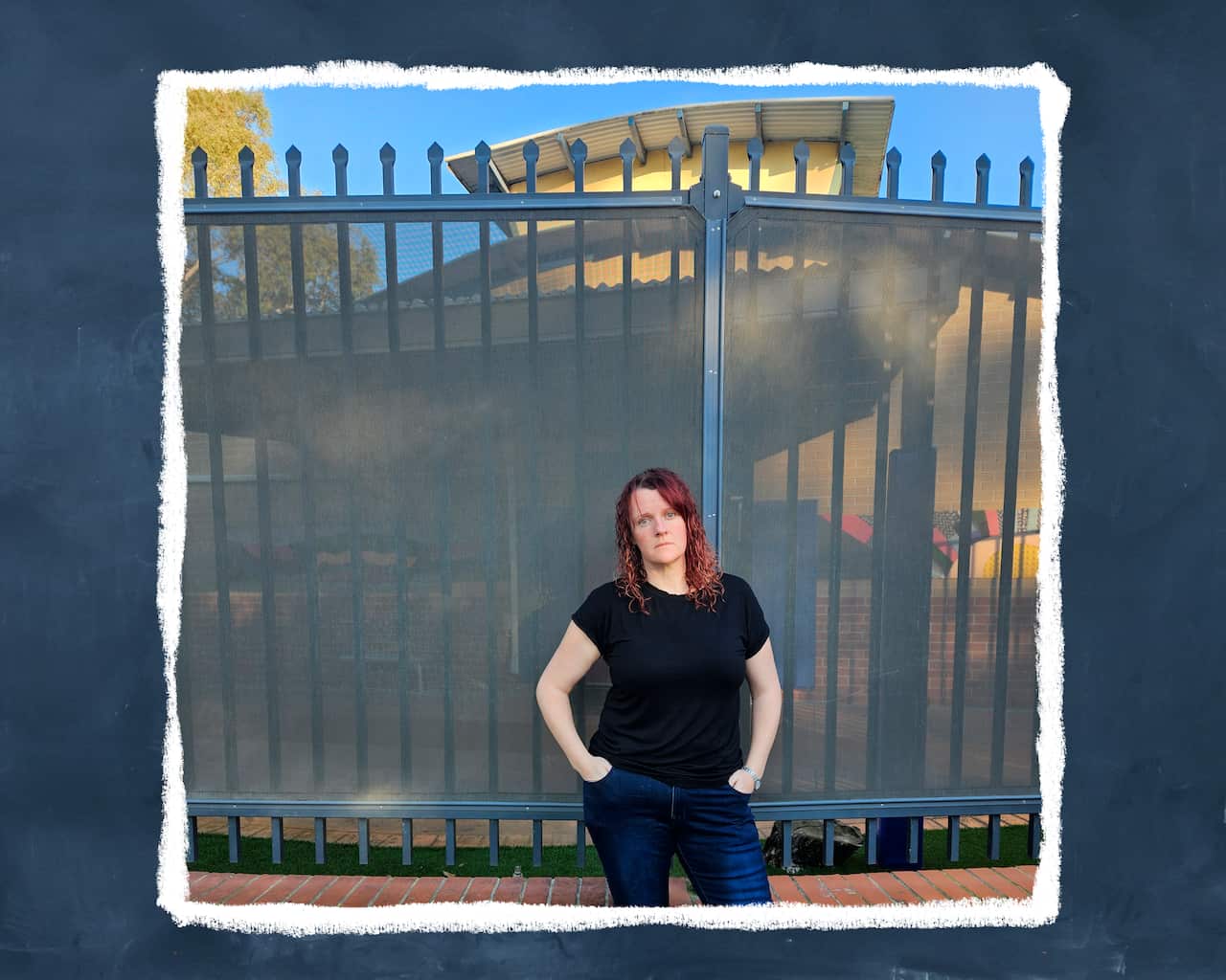 A woman with red hair wearing a black T-shirt stands in front of a fenced outdoor area of a school. 
