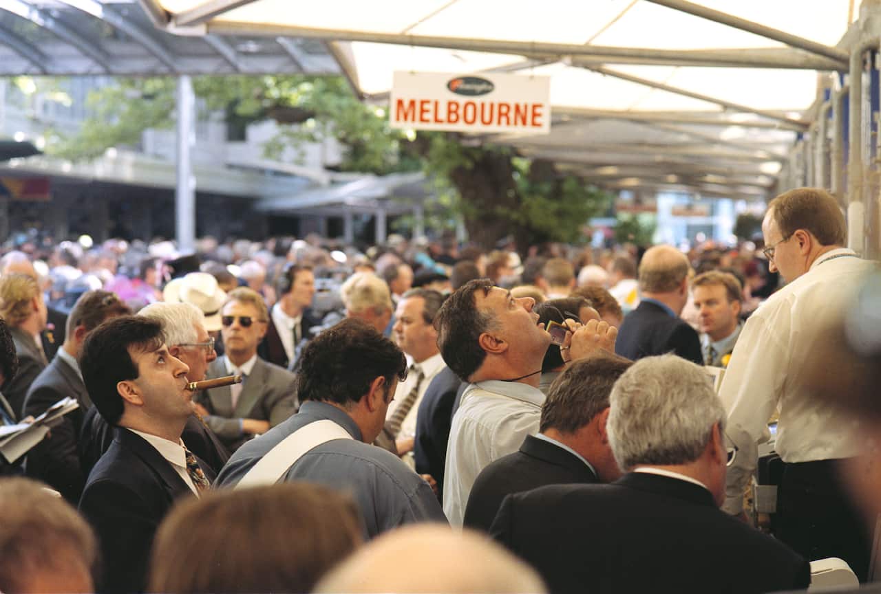 A group of men in suits gambling at the Melbourne Cup in 2000. They are watching a television screen.