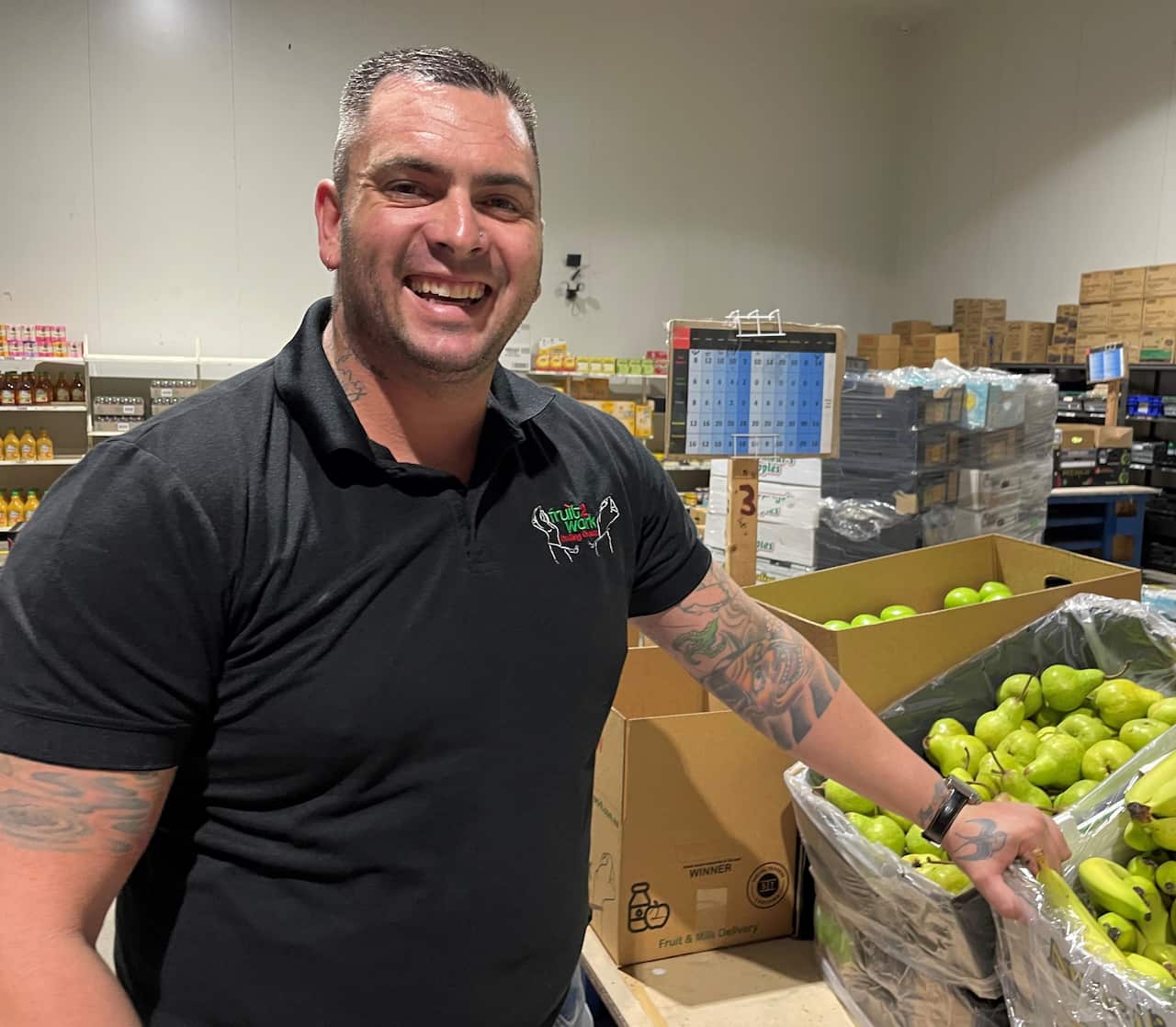 A man in a black t-shirt standing next to a box of pears in a warehouse.