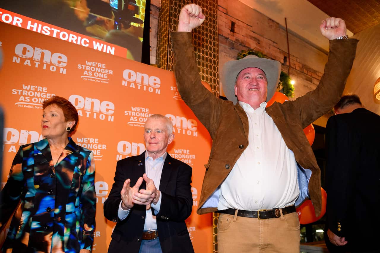 A white man wearing a brown jacket and akubra-style hat raising his arms in celebration. A man and a woman stand beside him.
