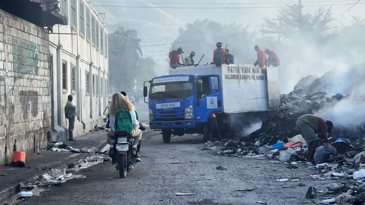 A scene of chaos and destruction in a city: a damaged building on the left and a truck next to a large pile of rubbish on the right. A woman with a green backpack is seen from behind, sitting on a motorbike behind a man. 