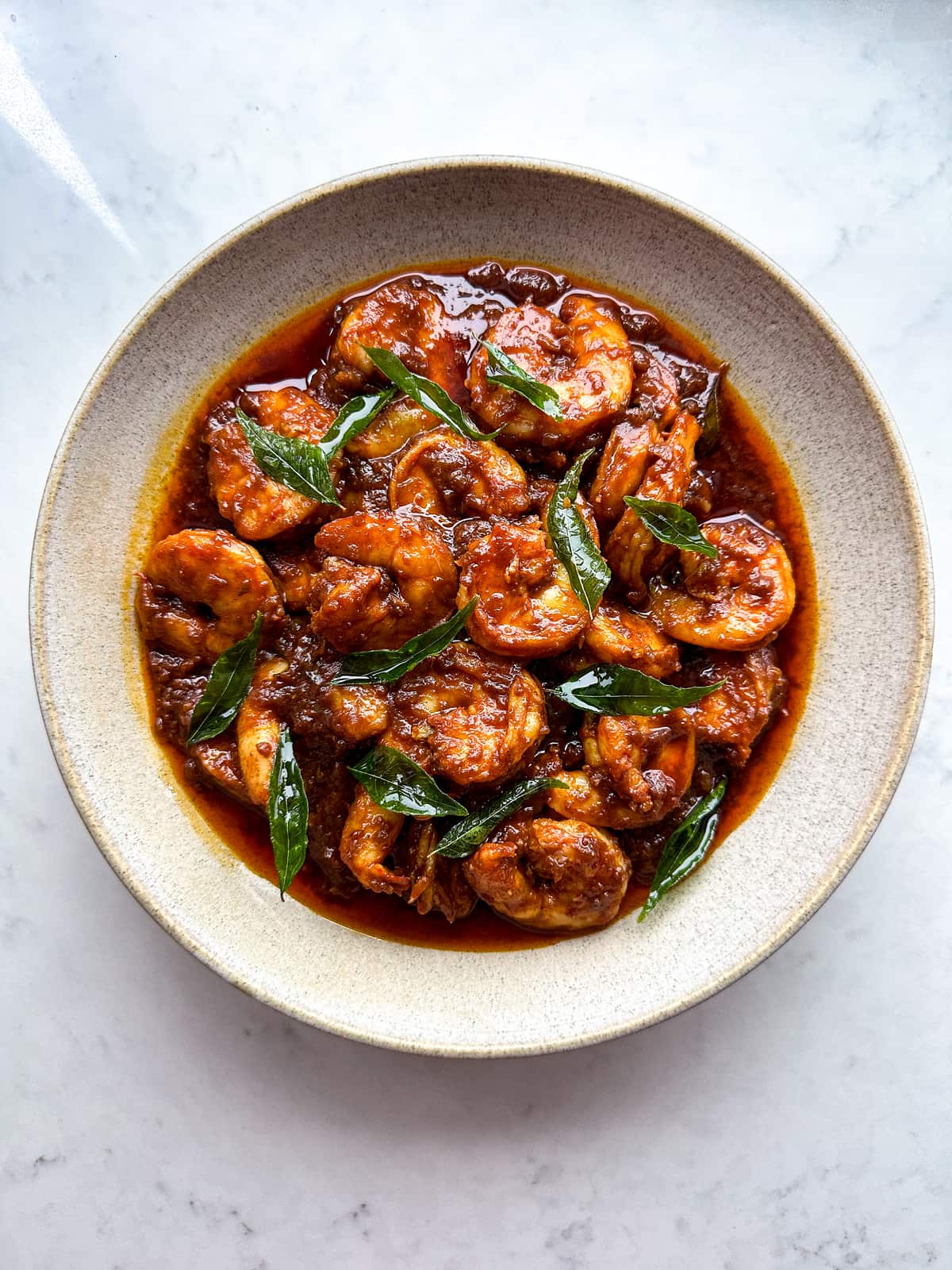 Prawns in a dark brown sauce or curry topped with curry leaves in a bowl.