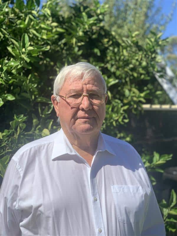 A man in a white shirt stands in a garden with a serious expression on his face.