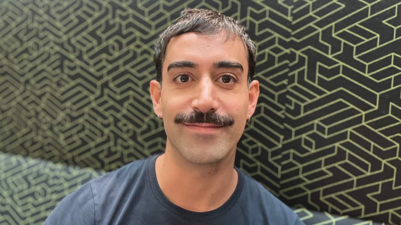 a closeup of a young man with a dark moustache and sleeper earring on a patterned couch