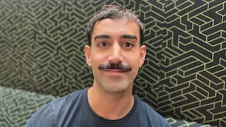 a closeup of a young man with a dark moustache and sleeper earring on a patterned couch