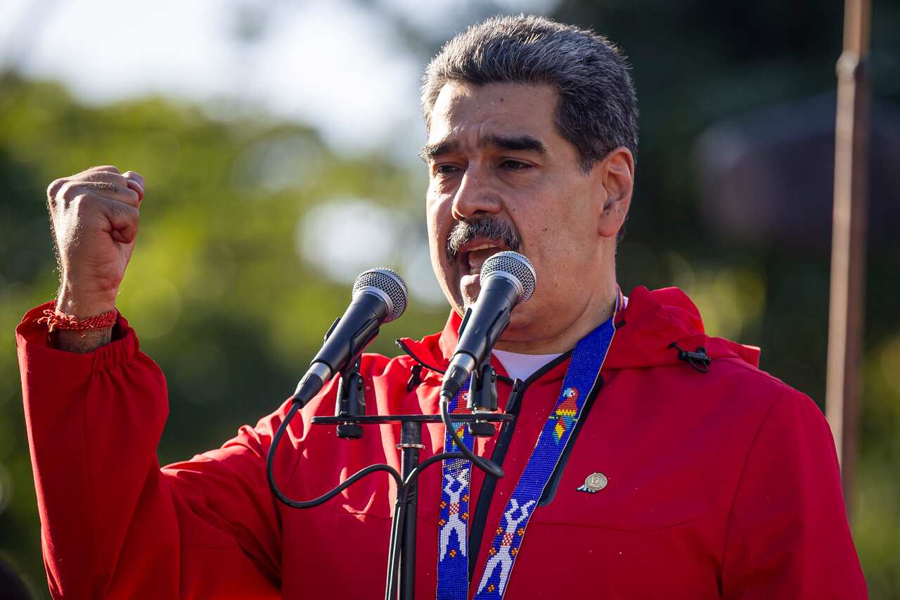 A man in a red jacket and a thick moustache closes his hand in a fist as he speaks into microphones.