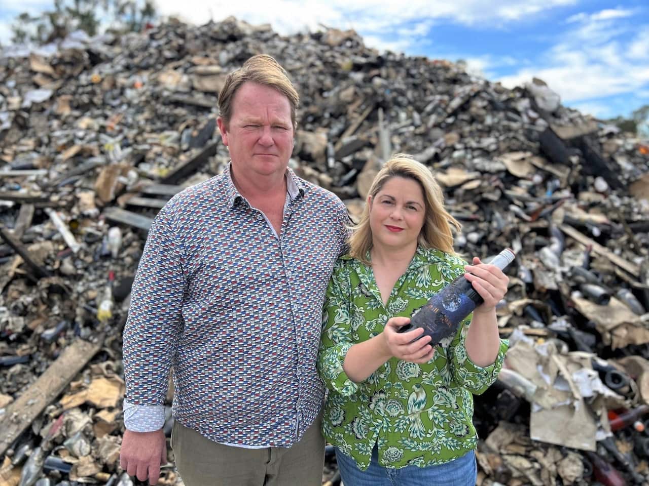 A man in a checked shirt stands next to a woman in a green shirt who is holdng a sparkling wine bottle, in front of a mound of debris.