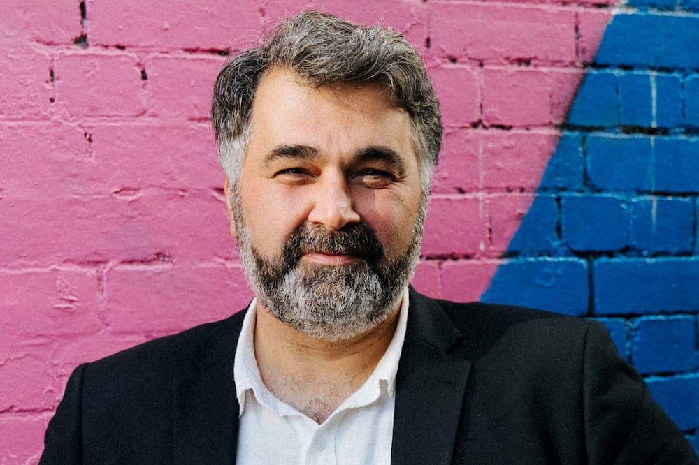 Kon Karapanagiotidis, wearing a white shirt and black blazer, standing in front of a pink and blue wall.