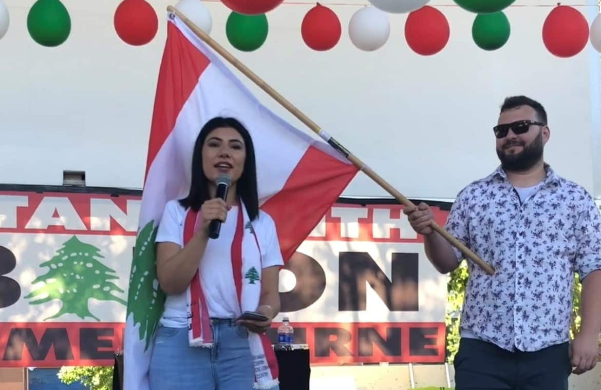 Fatima Hoblos is holding a microphone speaking on stage in front of a Lebanese flag.
