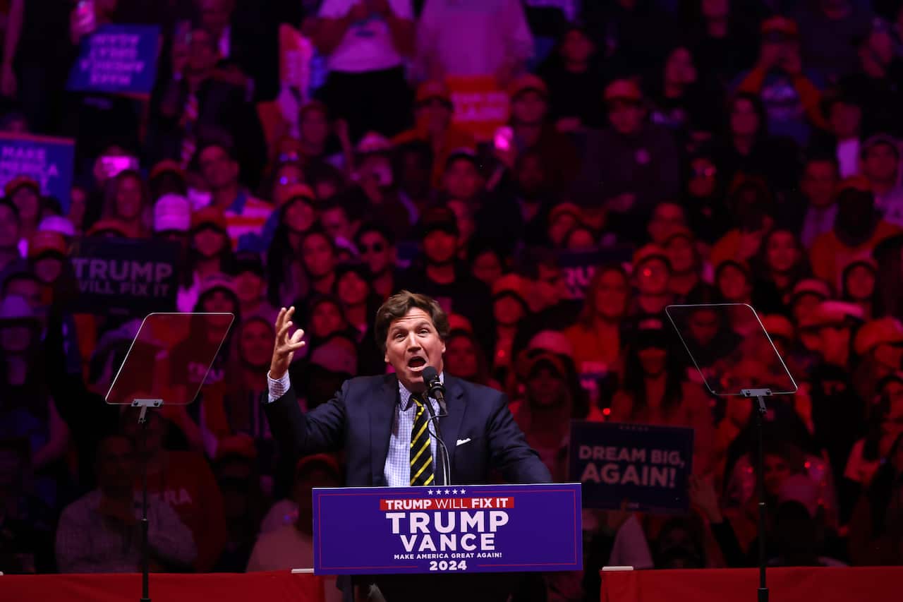 Tucker Carlson speaking at a podium that says "Trump Vance" on it. A crowd is seated behind him.