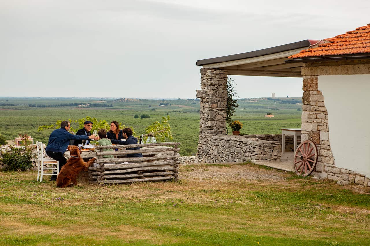 A small group of people sit at a table near a stone building. A green field stretches to the horizon behind them. A dog sits beside the table. 