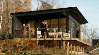 Two men are having their photo taken on a verandah of a wooden house by a woman in a long brown coat