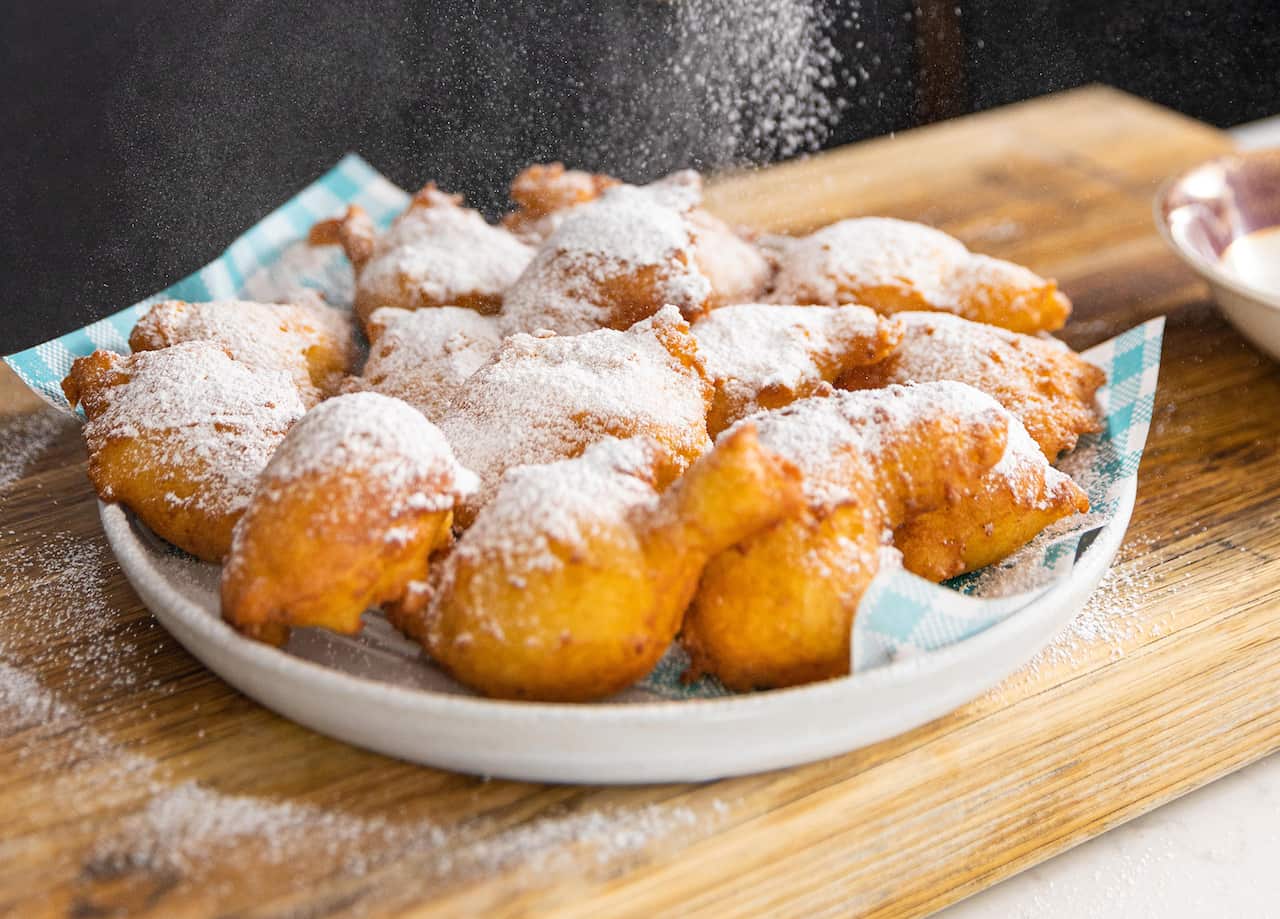 A white plate with a checked blue liner holds a layer of golden fritters, dusted wiht icing sugar. 