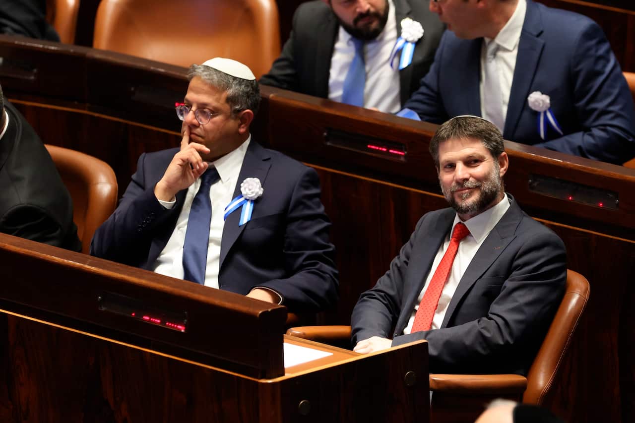 Two politicians, dressed in suits, sit in chairs in front of a long wooden table.