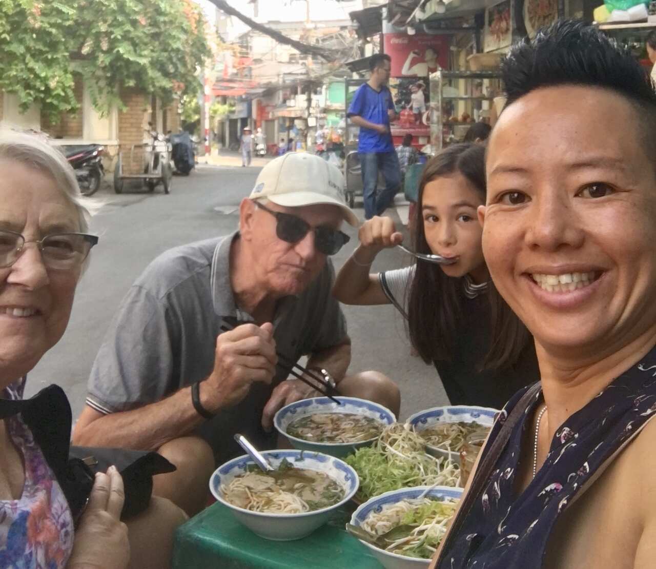 A middle-aged woman, a young girl and two older people sit hunched over a plastic table, eating bowls of Vietnamese noodle soup.