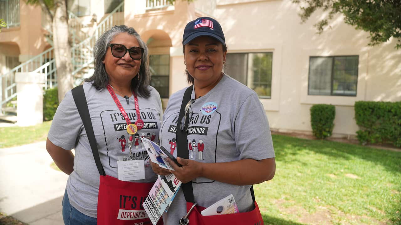 Two women are wearing grey t-shirts and red crossbody satchels, with one of them holding pamphlets.