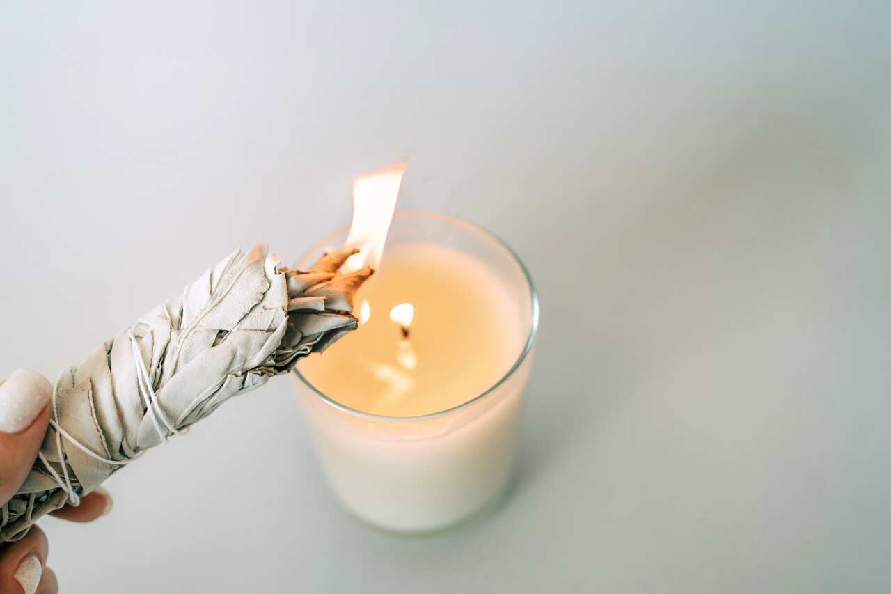 Woman is holding in a hand and set fire White Sage above burning candle on gray background. Set for aromatherapy and rituals. Top view