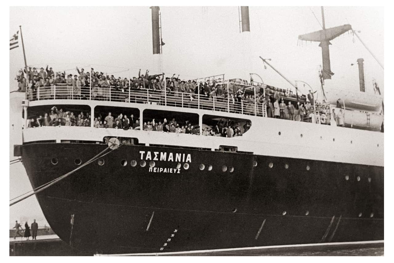 A black-and-white archival photograph of the passenger ship Tasmania docked in port, with its name written in Greek on the hull. Hundreds of passengers crowd the ship's upper decks and lean over the railings, waving toward the camera. A Greek flag flies at the stern, while cranes and other port equipment surround the vessel.