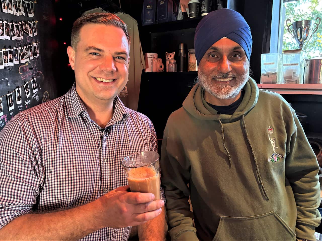 A man in a checkered shirt stands at a bar holding a glass of tea next to a man in a green sweater and a blue turban. 