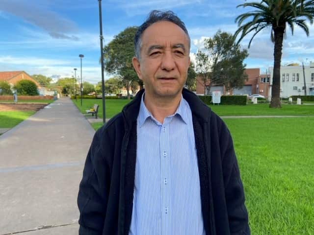Mehmet Obul stands in a shirt and jacket in a public park. 