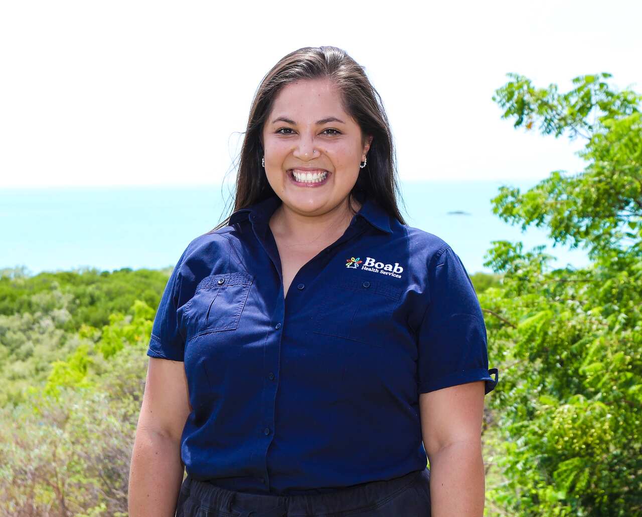 A woman in a blue shirt standing outside and smiling. The ocean is in the background.