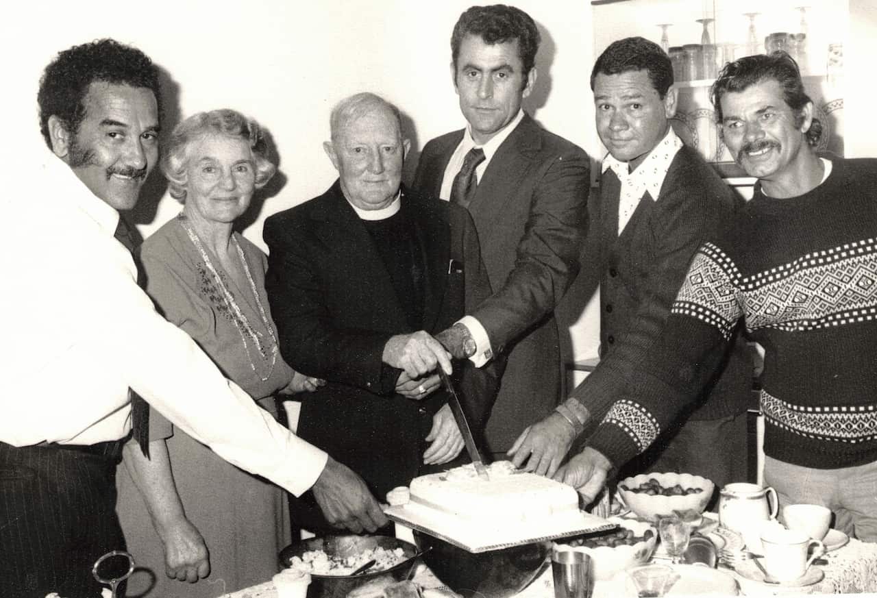 An elderly couple cuts a cake and poses with their male students who are now adults