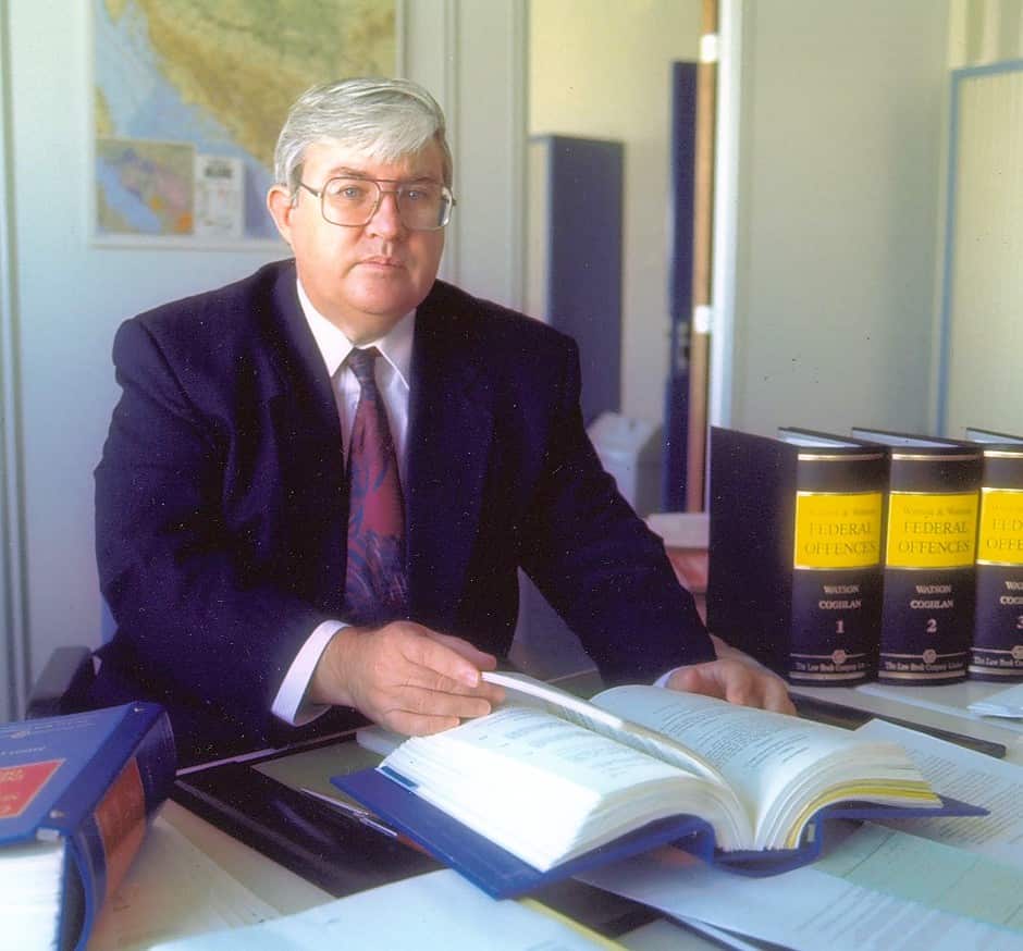 A middle-aged man with greyish white hair, wearing large rimmed glasses, looking at the camera. He's wearing a dark suit, with a white shirt and a red and purple swirled tie. He's sitting at an office desk, flicking through a thick legal book. More legal books are beside him, and a map is on the wall behind him.