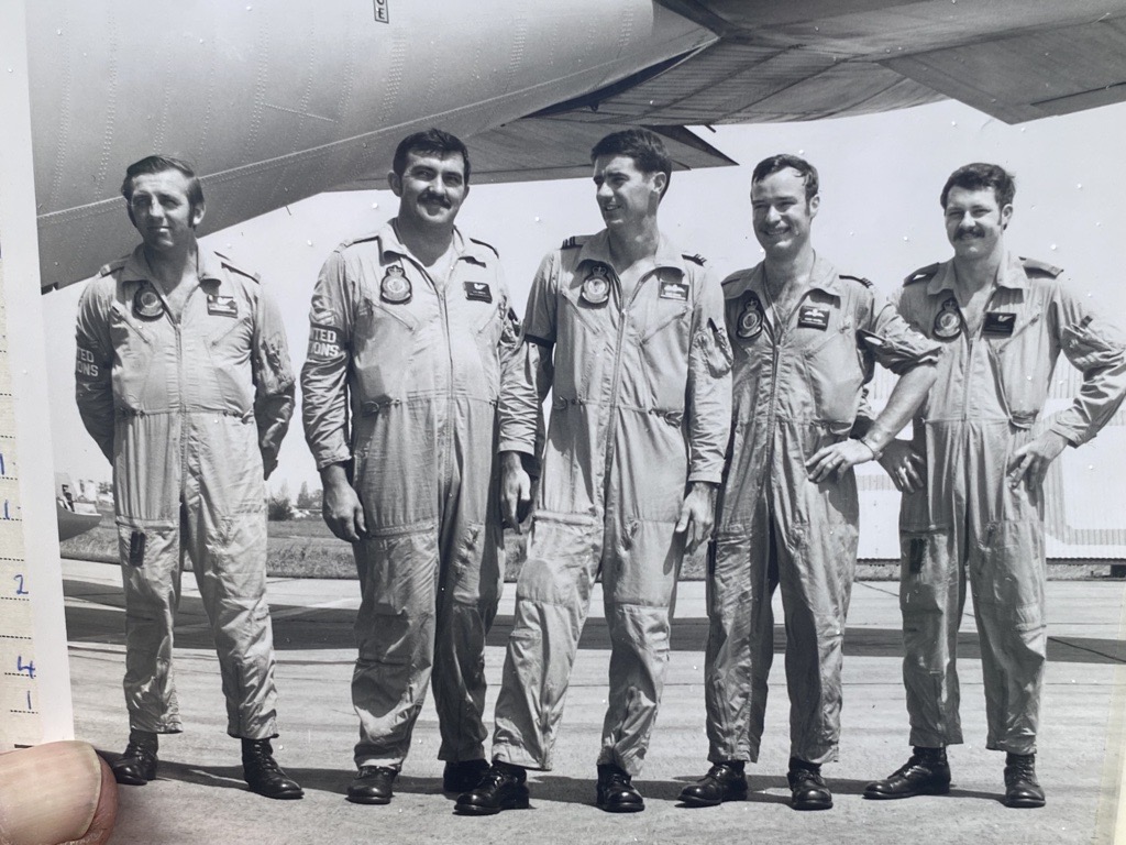 Old black and white photo of five pilots in overalls standing side by side, with an aircraft behind them.