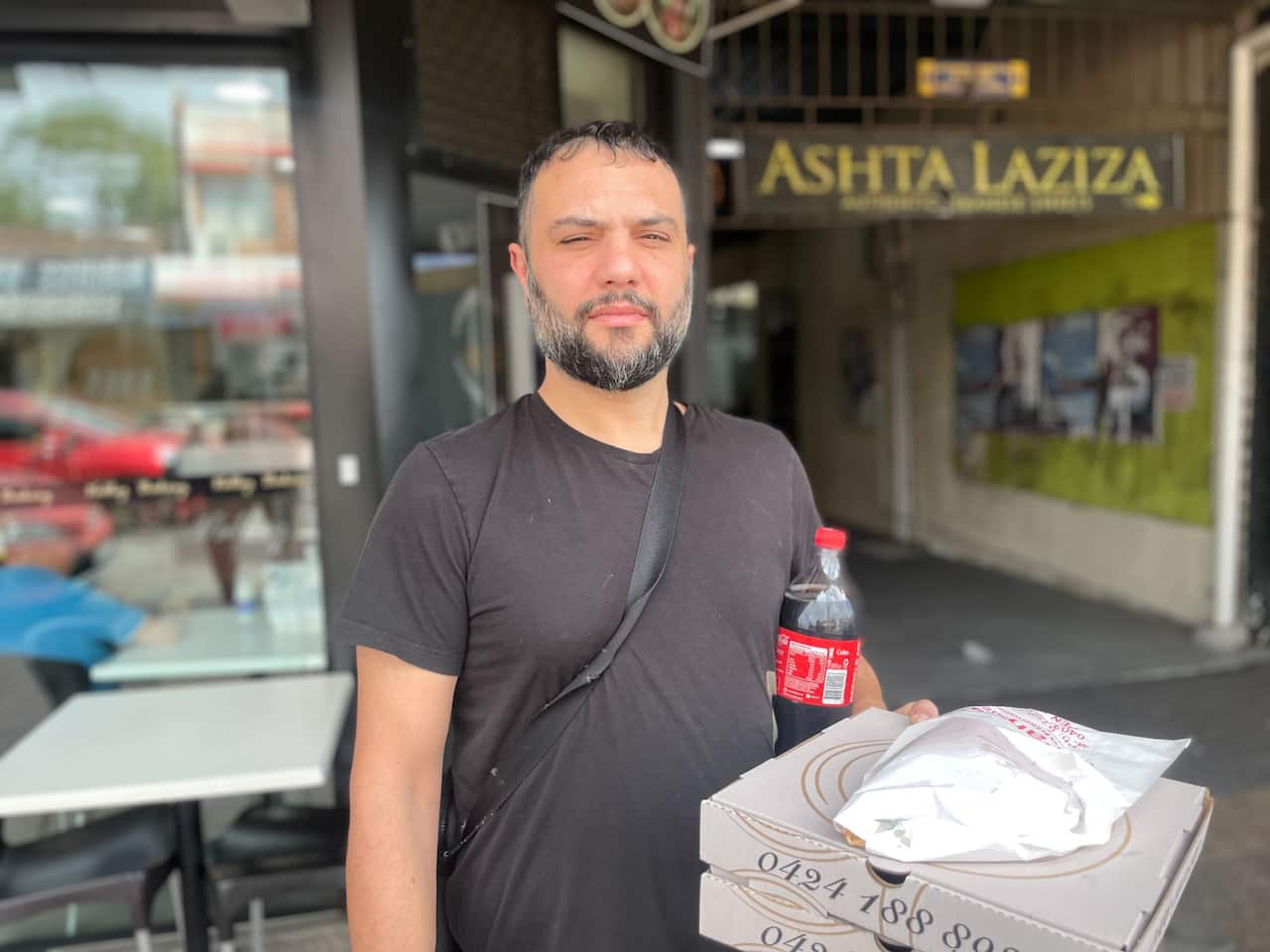 A man in a black t-shirt holds a bottle of Coke and two pizzas. 