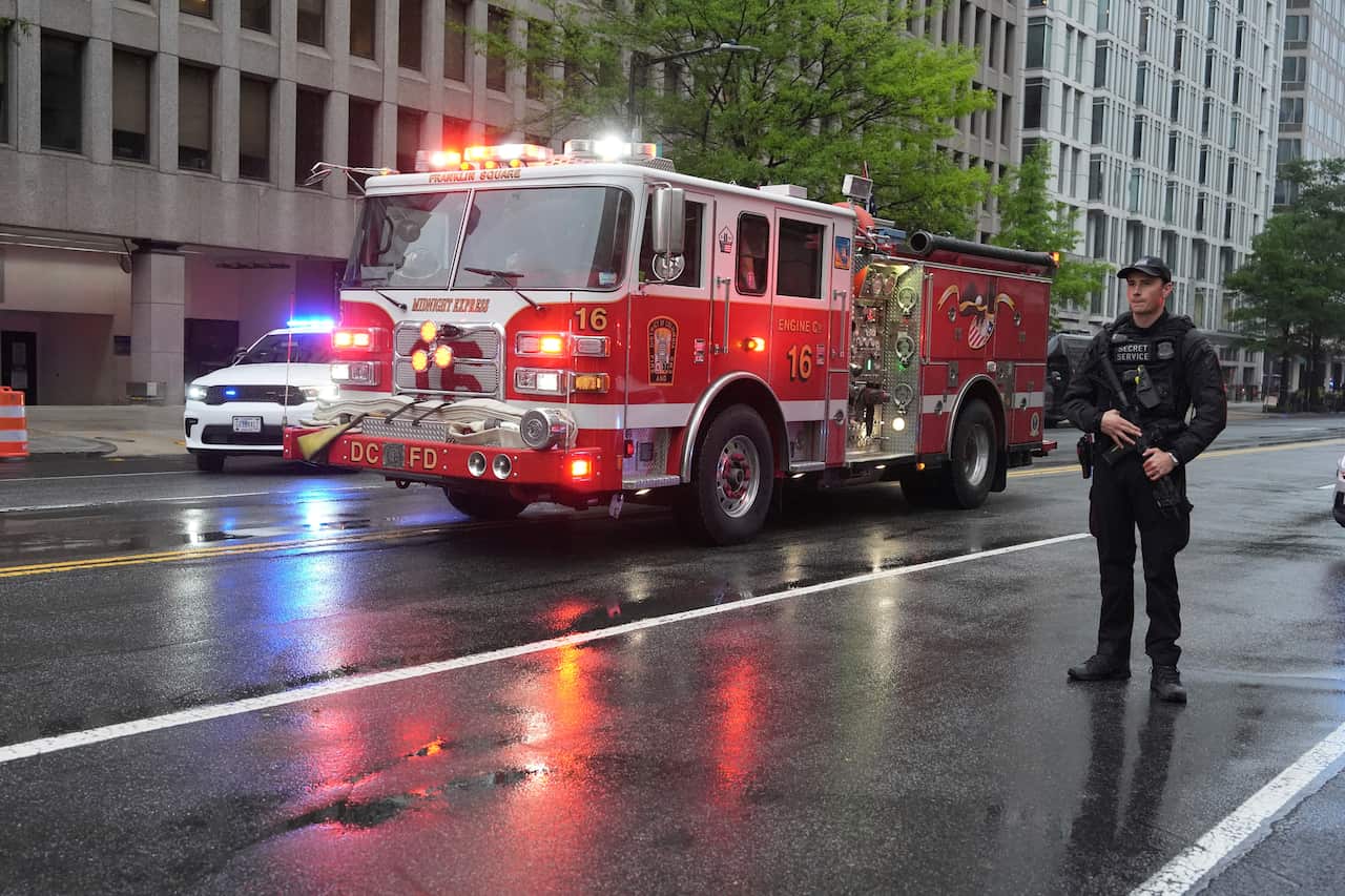 A fire truck with its lights on is driving in the street. There is a Secret Service agent standing.