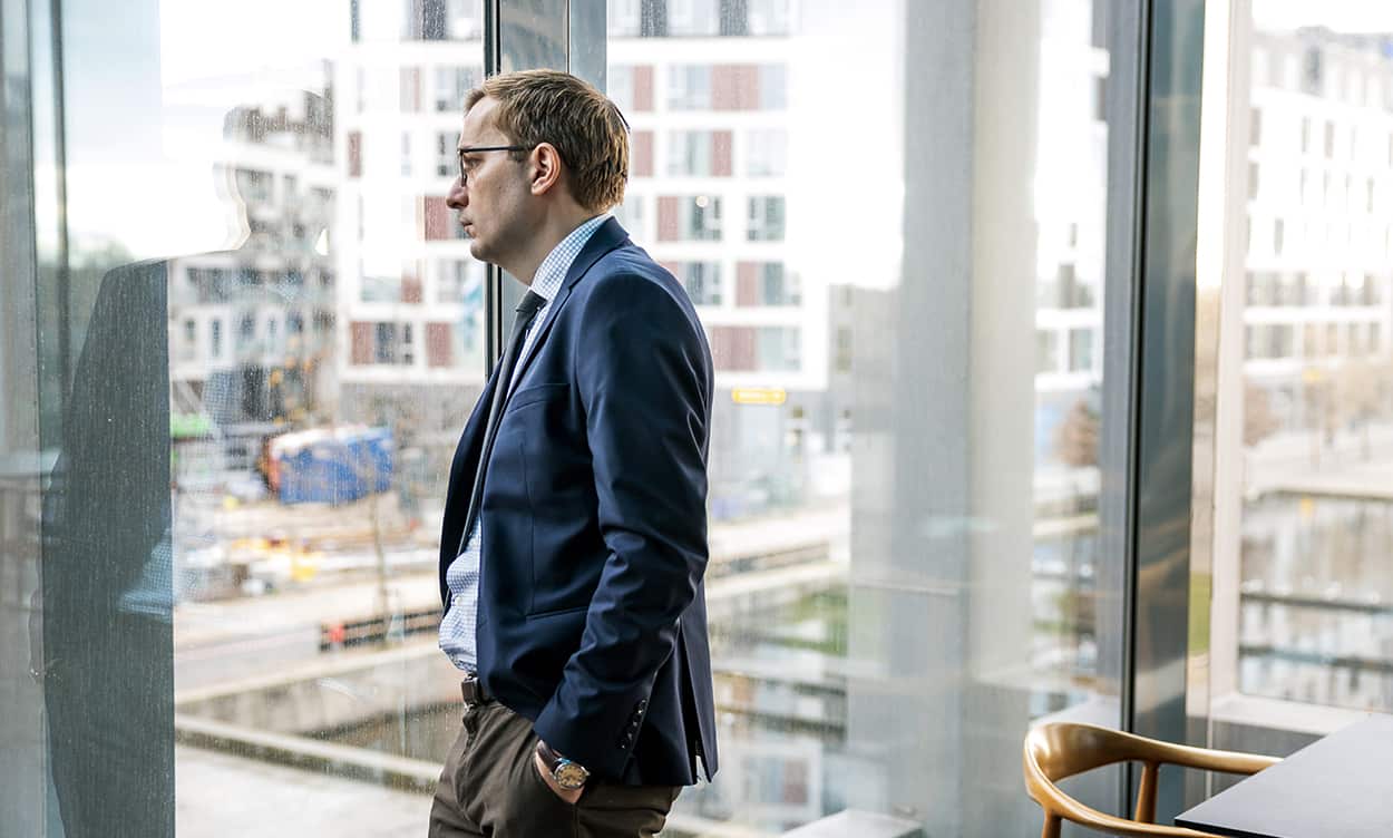 A man in a blue business suit stands looking pensively out through full-lenth glass windows at a city scape.