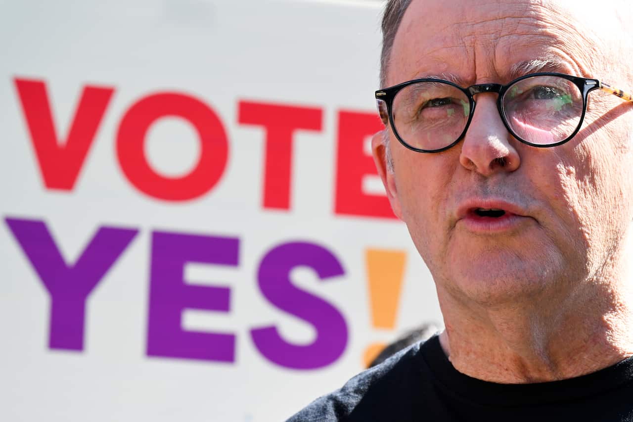Anthony Albanese, wearing a dark t-shirt and glasses, speaks in front of a sign reading Vote yes!