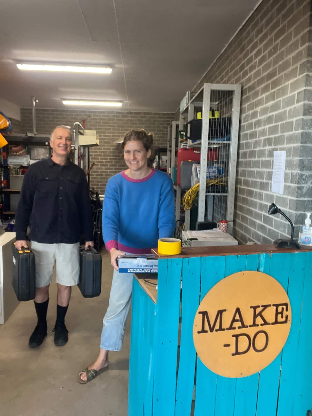 Volunteers, a man and a woman, man a desk which has a sign saying "Make-do' in a shed full of random tools and appliances.