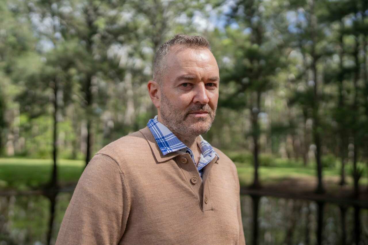 A middle-aged man with short grey hair wears a taupe sweater and stands in front of a pond in a park.
