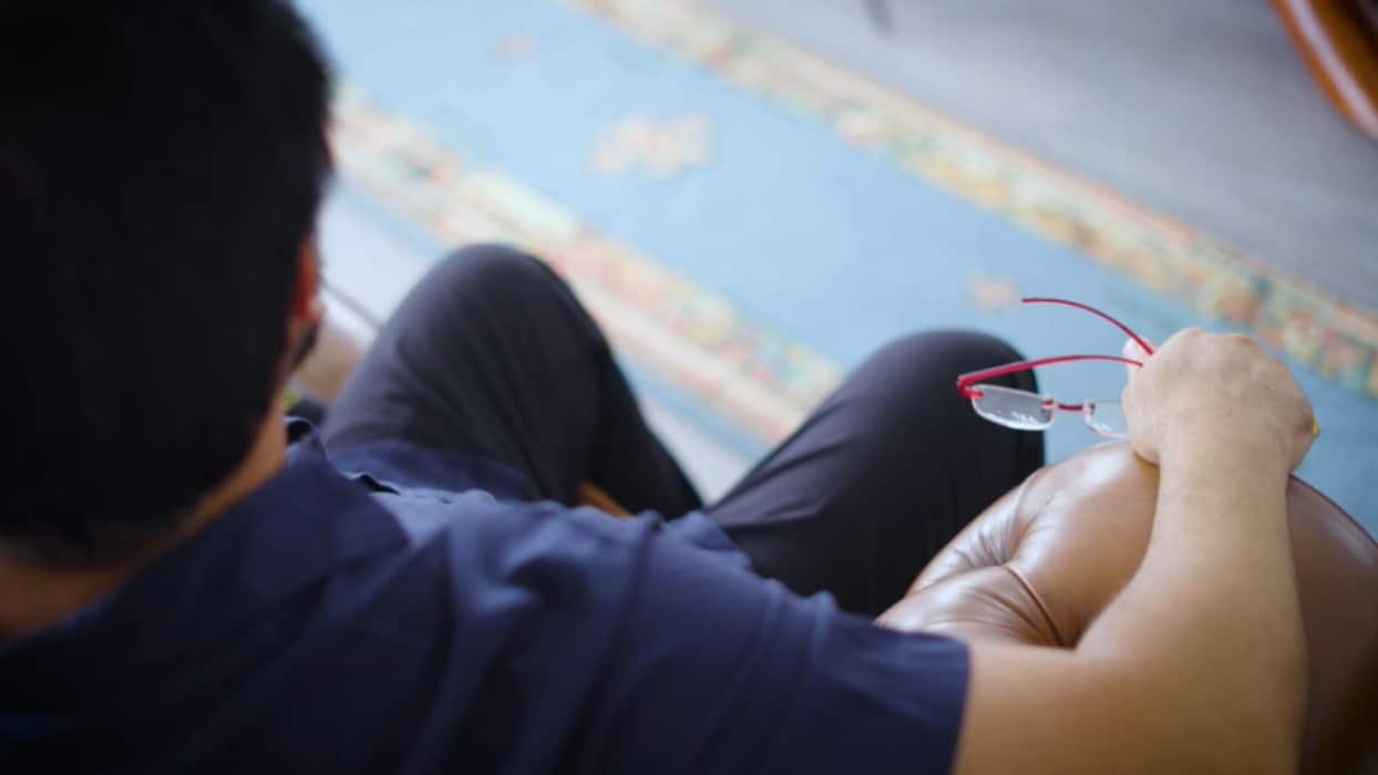 An anonymous picture of the back of a man's head and his lap. He is holding a pair of glasses in his hand. There is carpet in the background.