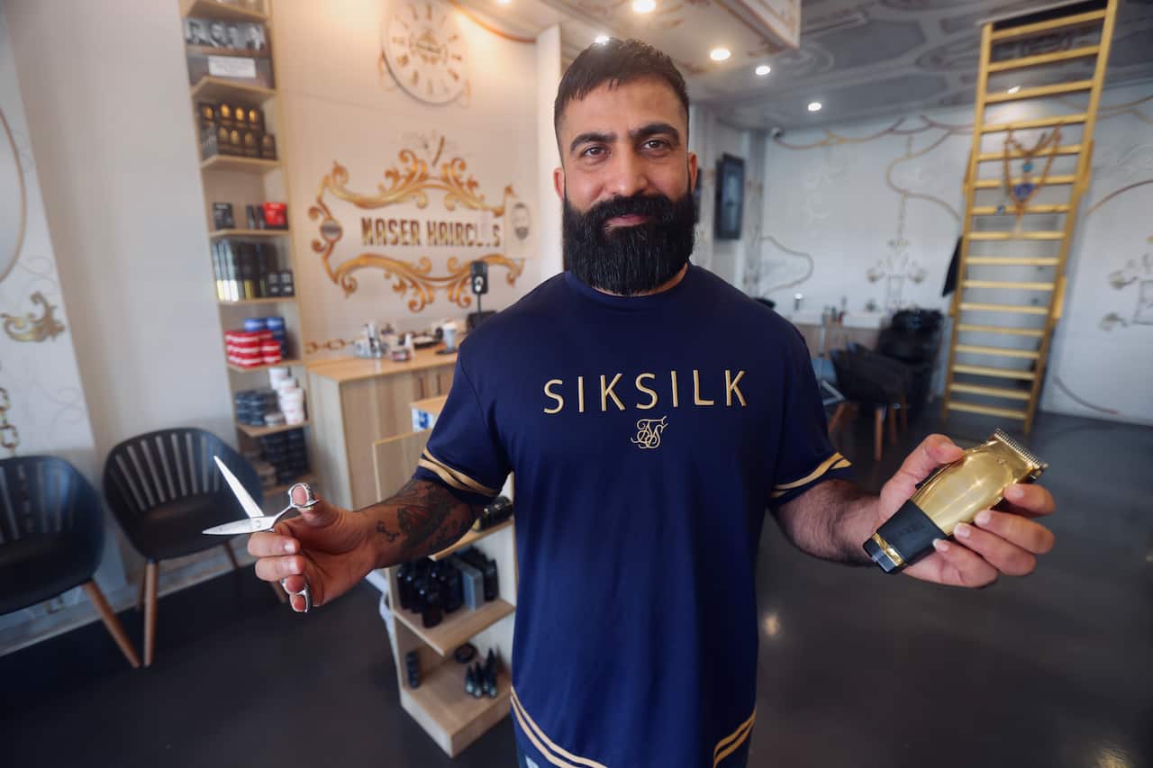 A barber with a black beard holding hair clippers and scissors, posing for a photo. He is wearing a dark blue t-shirt with the word Siksilk written across the chest in gold writing.