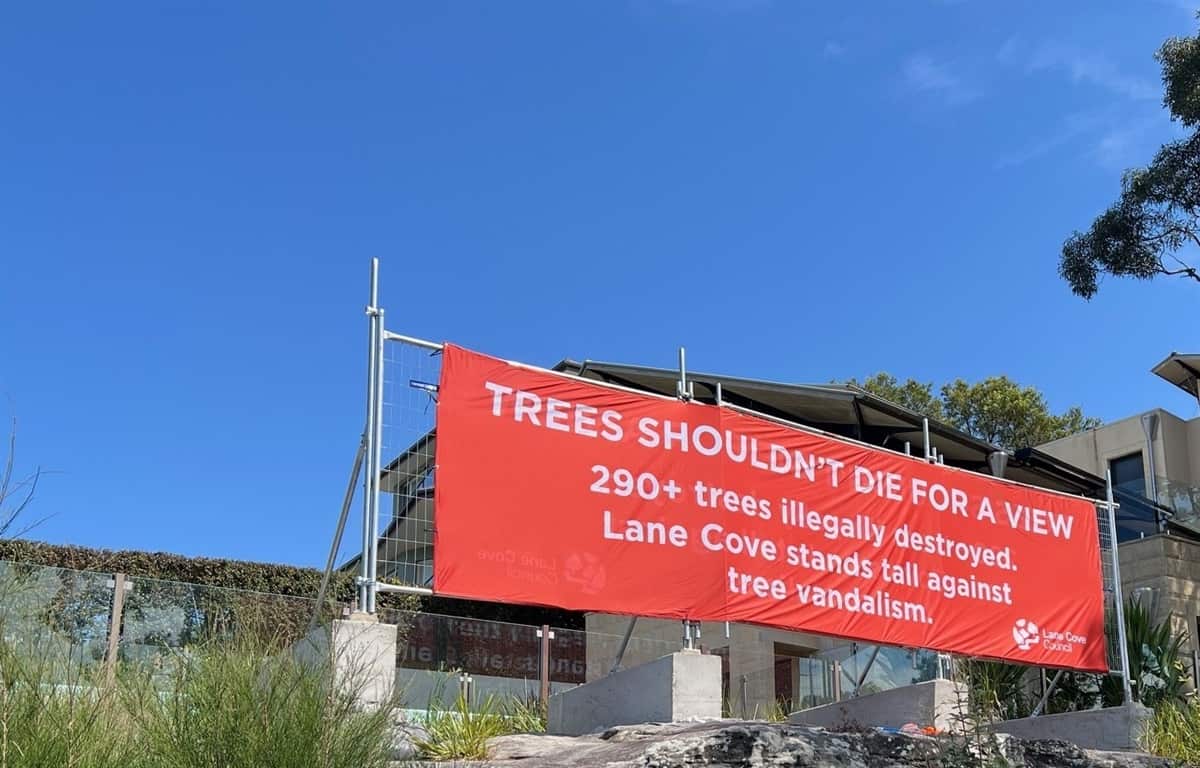 An orange banner hangs on a fence outside a house that reads: Trees shouldn't die for a view.