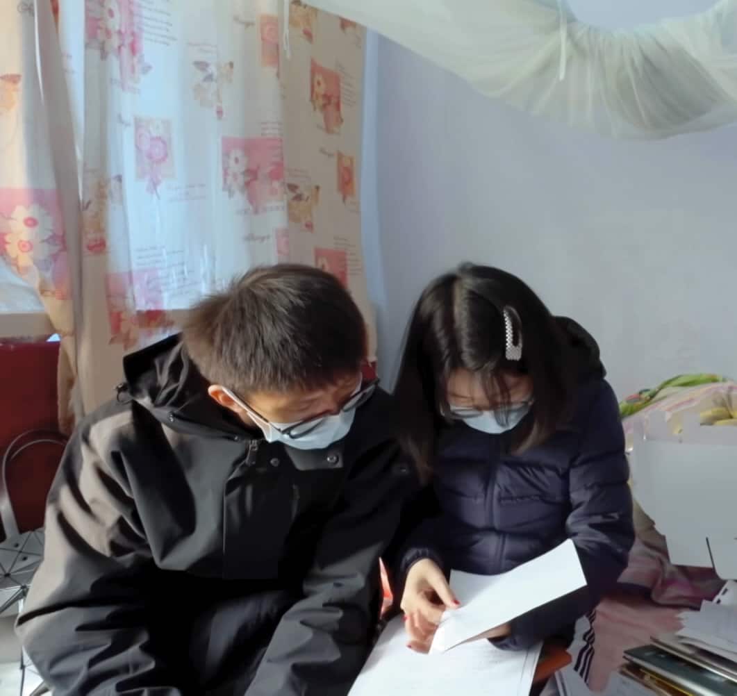 A man and woman sit on a bed looking through documents. They are both wearing glasses and face masks. 