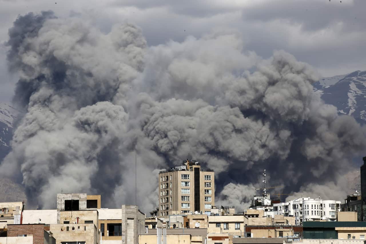Large plumes of thick gray smoke rise from behind a row of residential buildings and skyscrapers in an urban area, with snow-capped mountains visible in the distance under a cloudy sky.