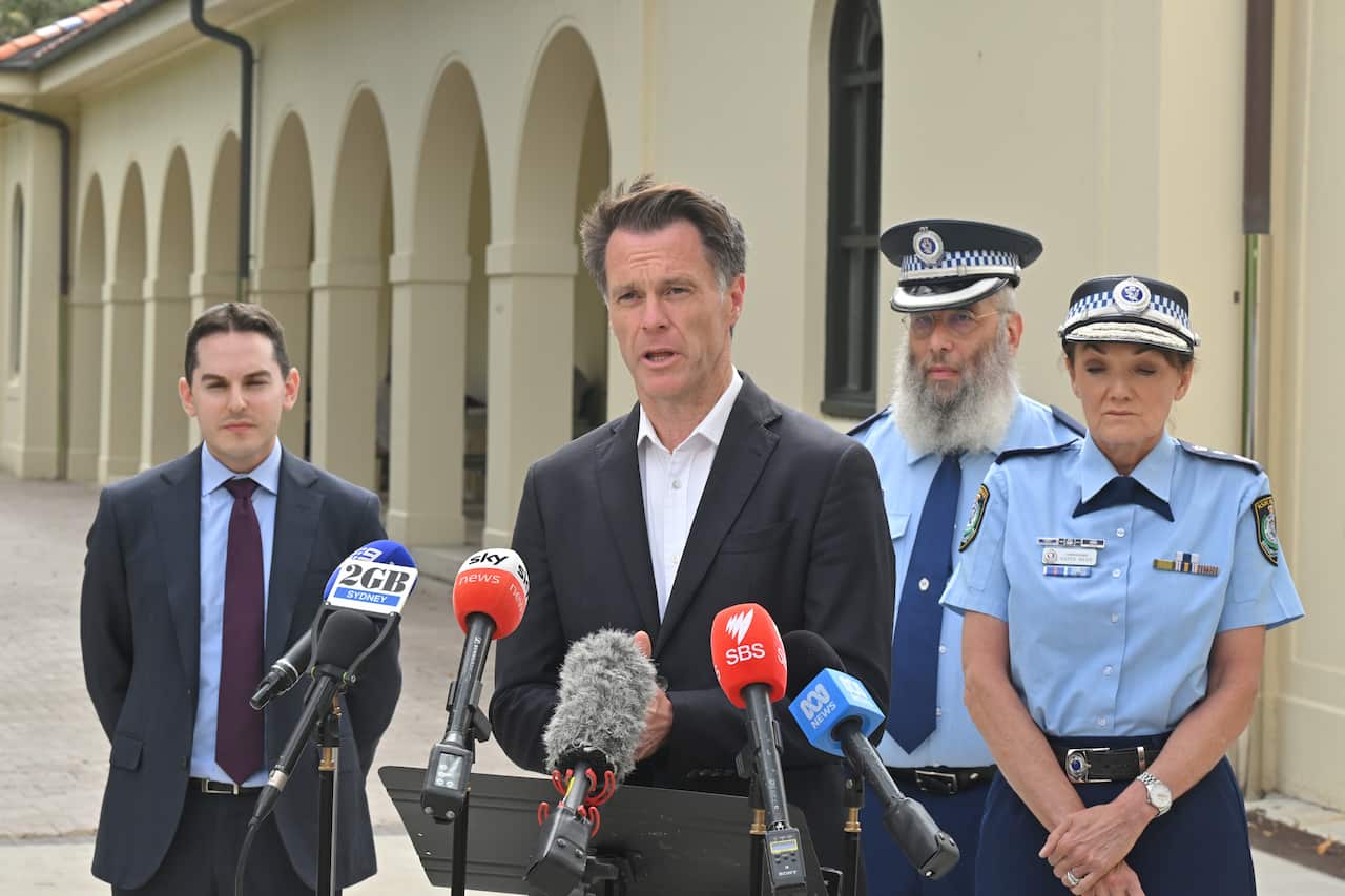 A man in a suit speaks into media microphones at a press conference outside. A male and female police officer in blue uniform stand behind him.