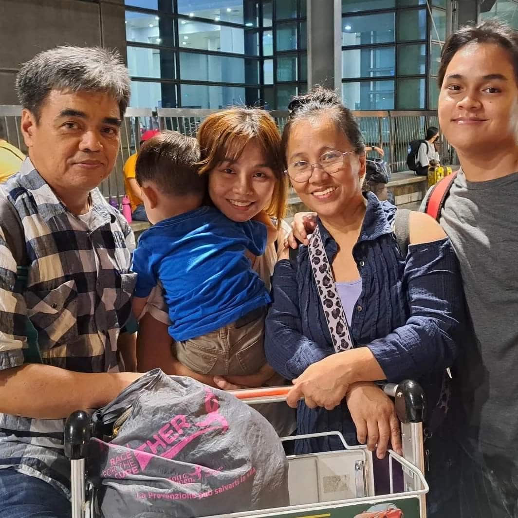 A group photo of a family standing in front of an airport trolley. From left to right: an older man in a black-and-white checkered shirt, a young woman holding a toddler in a blue t-shirt, an older woman wearing reading glasses and a denim jacket, and a young man in a grey t-shirt. 