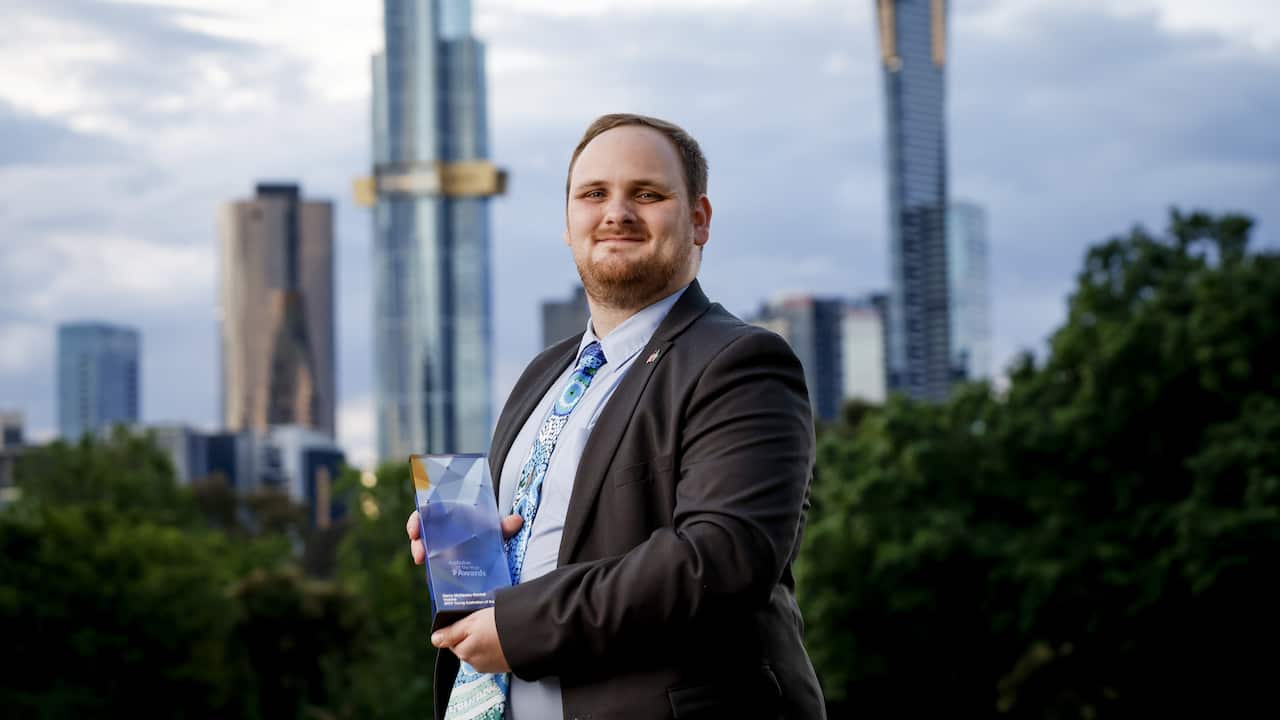 Darcy McGauley-Bartlett in a suit holding a trophy and standing outside