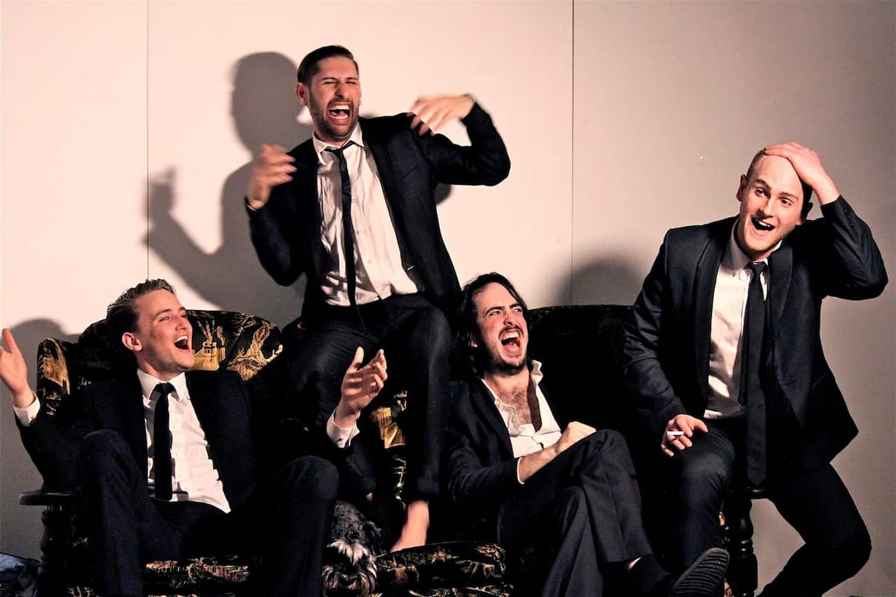 Four young men in black suits sitting on a couch and laughing