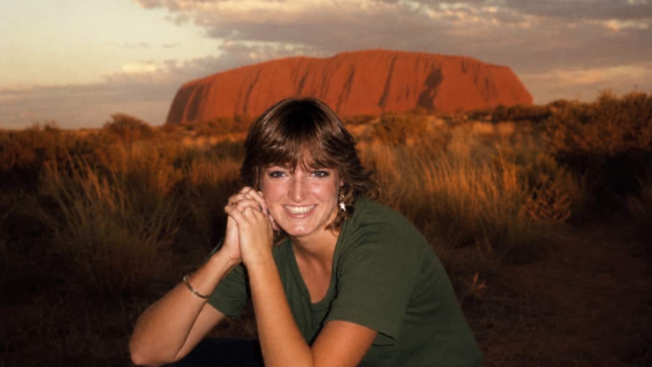 a young brunette woman smiles crouching down in front of Uluru at sunset