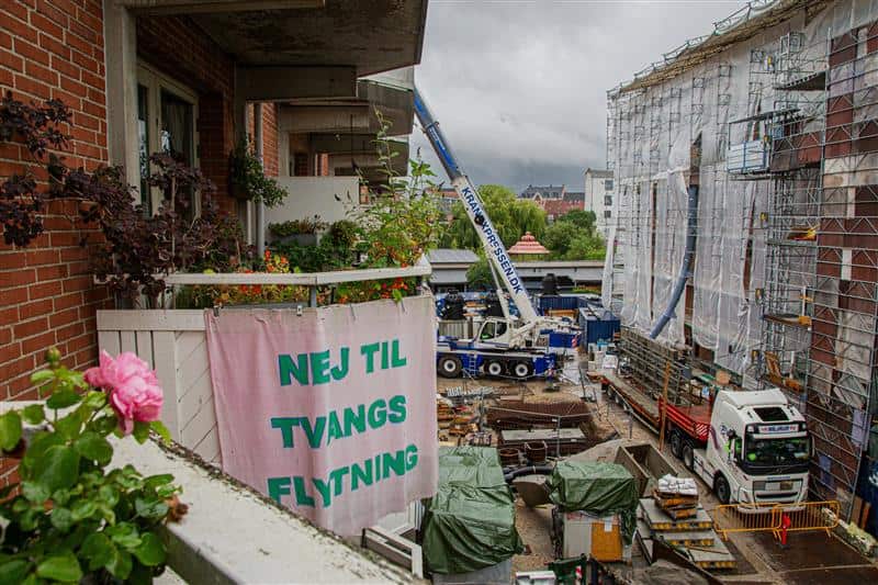 On the left, an apartment with a balcony full of plants has a pink sign reading 'no to forced relocation' in Danish. The rest of the image shows a truck and a crane on a building site below the apartment, with a building covered in scaffolding on the right. 