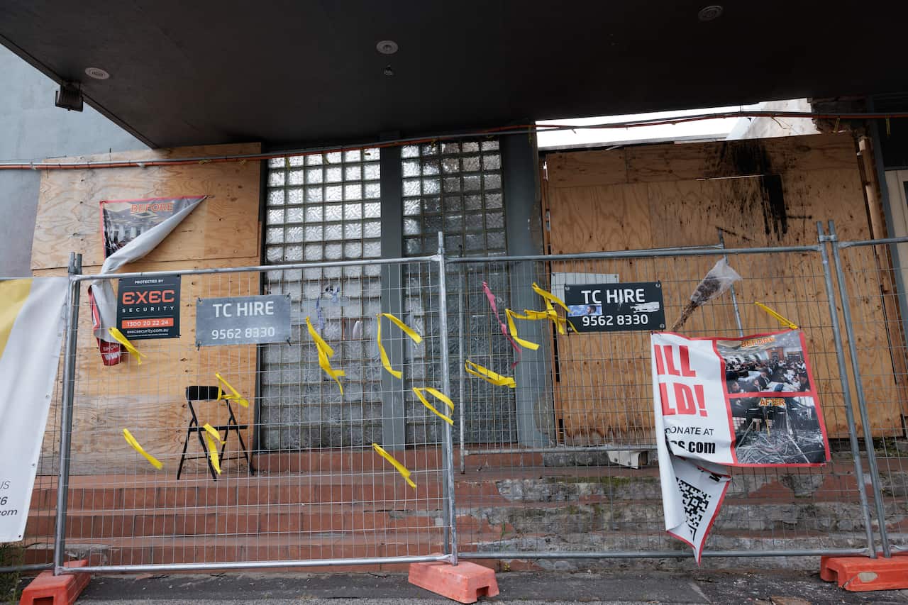 A building entrance boarded up with plywood and secured behind a metal fence featuring "TC Hire" signs and yellow caution ribbons.