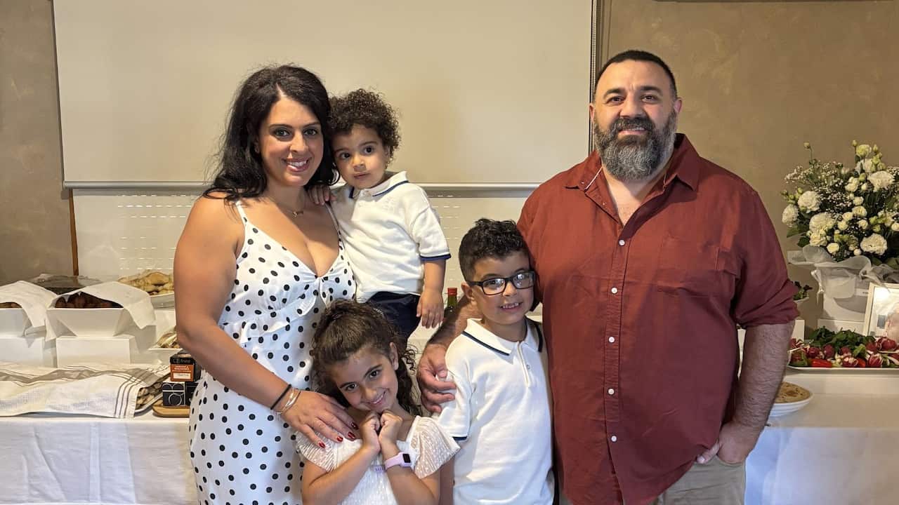 a young family - a mum, dad and three young children - smiling in front of a white table with a spread of food in it